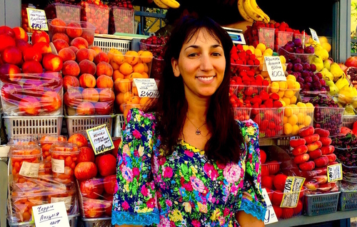 Behind Emily is a stand containing fruits from Central Asia. Her research aims in part to untangle the myriad ways both the fruits and the people selling them come to Saint Petersburg.